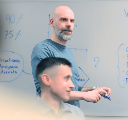 Foto fr&aring;n en l&auml;rarledd utbildning i 2c8 Apps d&auml;r en man st&aring;r framf&ouml;r en whiteboard och f&ouml;rklarar n&aring;got f&ouml;r en grupp m&auml;nniskor.